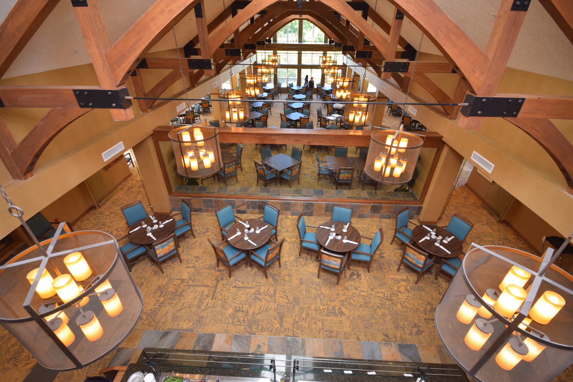 Interior view of a dining room with multiple round tables and blue cushioned chairs arranged neatly. The room features large wooden beams on the ceiling and modern hanging light fixtures. A large mirror on one wall reflects the tables and chairs, enhancing the spacious feel of the room.