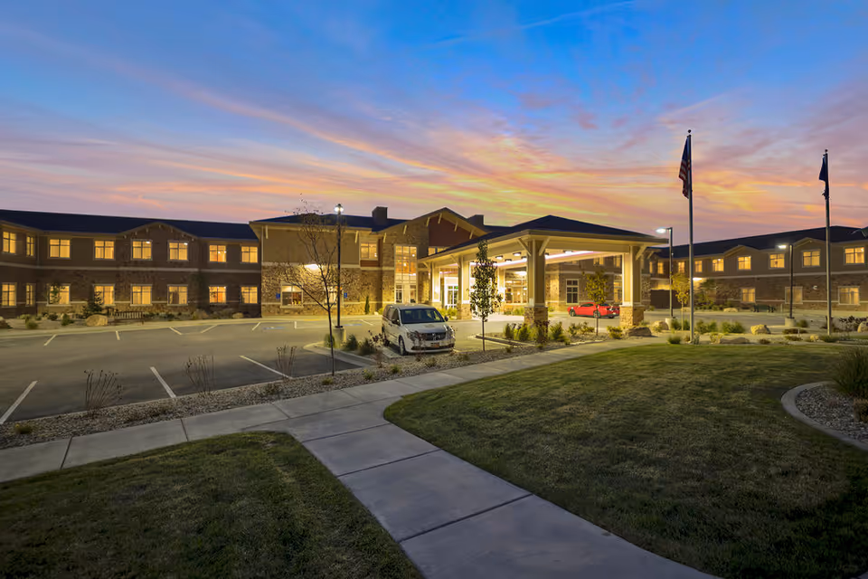 Exterior view of Mission At Crescent Senior Living facility at dusk, showing a large two-story building with many lit windows, a covered entrance with pillars, a parking lot with a few cars, landscaped greenery, and two flagpoles with flags.