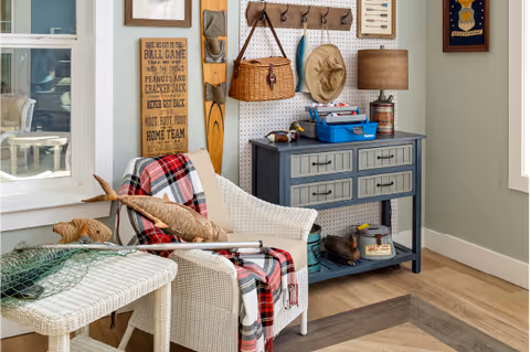 Cozy indoor sitting nook with wicker chairs draped in a plaid blanket, a blue console table with drawers, and fishing-themed decor on the wall.