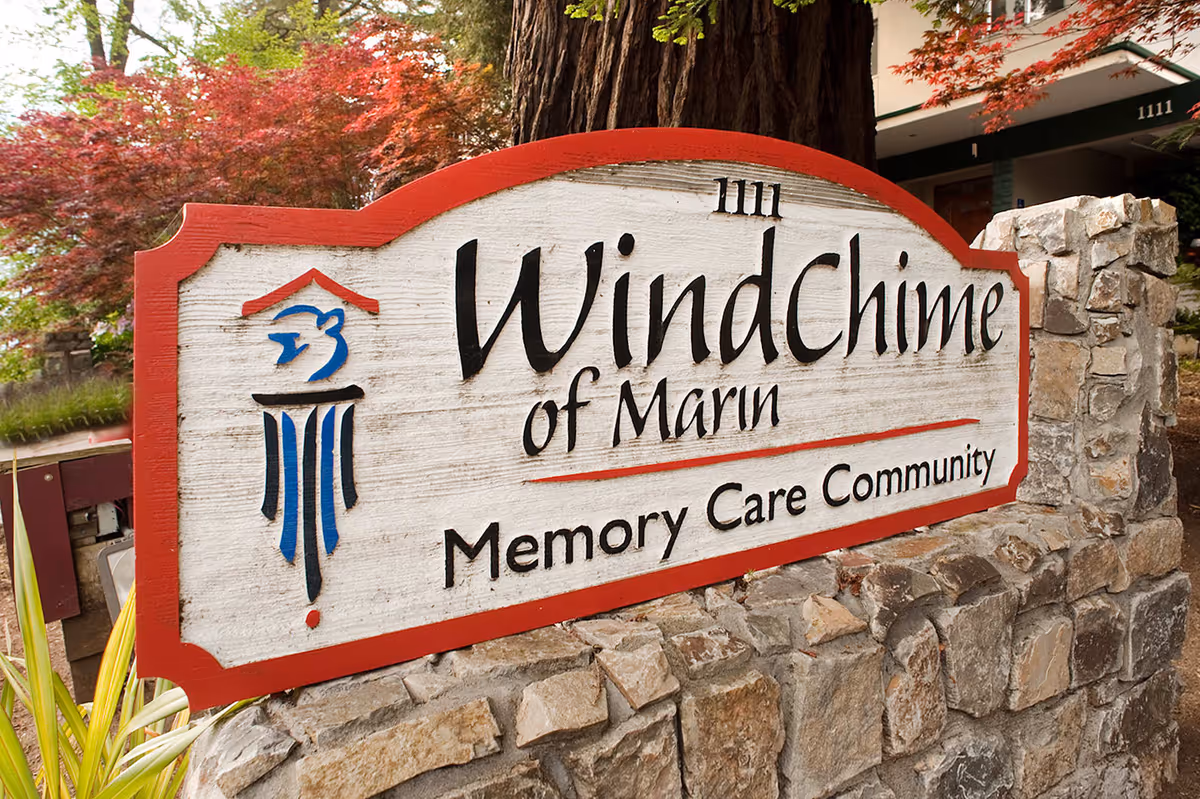 A wooden sign with red trim mounted on a stone base, displaying the text '1111 WindChime of Marin Memory Care Community' with a blue and red logo on the left side. The background includes trees with red leaves and part of a building.