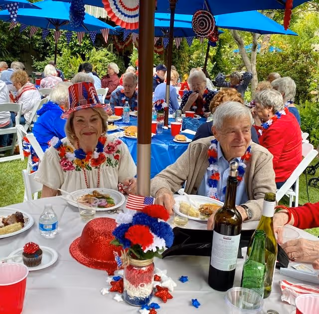A group of elderly people enjoying an outdoor patriotic-themed party with red, white, and blue decorations. They are seated at tables covered with blue and white tablecloths under blue umbrellas. One woman is wearing a red, white, and blue hat and a floral necklace, while others are also adorned with patriotic accessories. Plates of food, drinks, and festive centerpieces are on the tables.