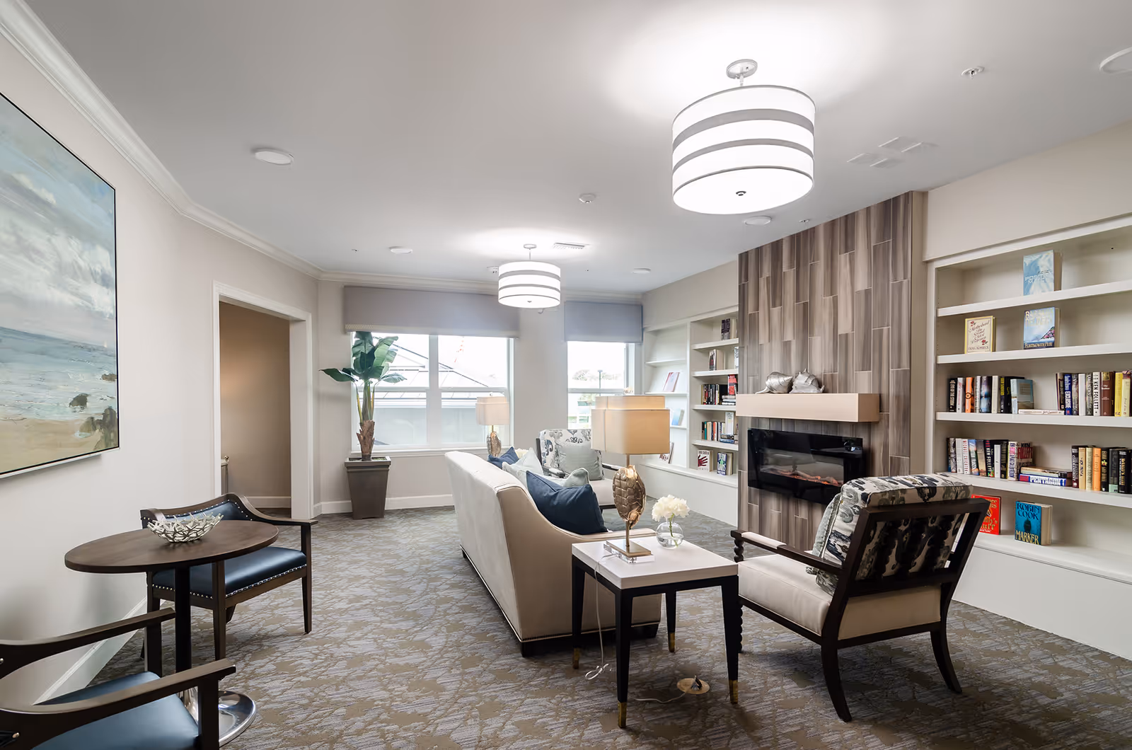 A bright and cozy living room area in a senior living facility featuring a beige sofa with blue and green pillows, two armchairs with patterned cushions, a small side table with a lamp and flowers, built-in bookshelves filled with books, a modern electric fireplace with a wood-paneled surround, large windows with gray valances, a potted plant, and two large ceiling lights.