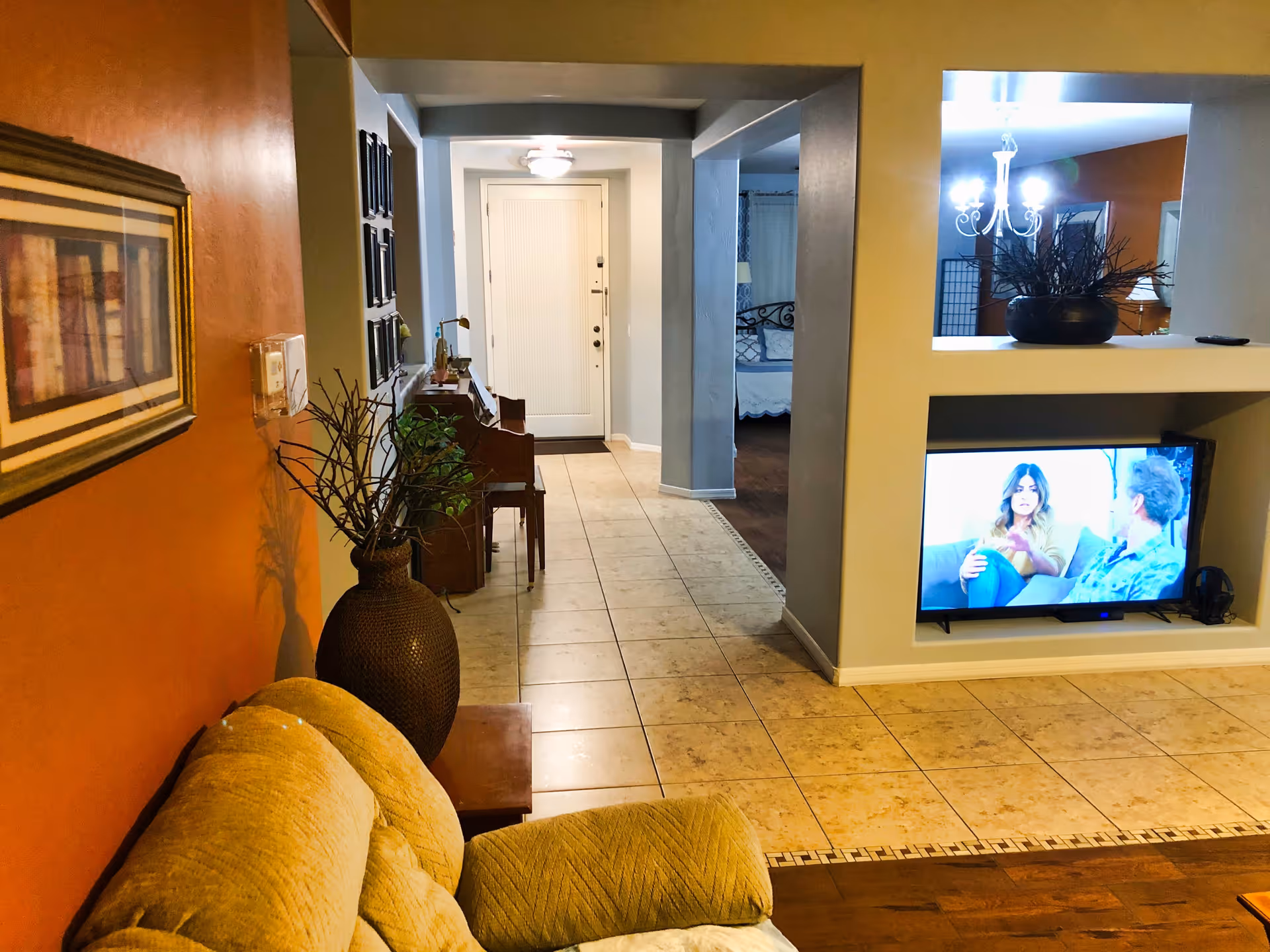 Interior view of a living space in Radiant Care Home showing a beige couch in the foreground, a large vase with decorative branches, a hallway leading to a white door, and a built-in wall unit with a television displaying two people having a conversation. The room has tiled flooring and warm-colored walls with framed artwork and a chandelier visible in the adjacent room.