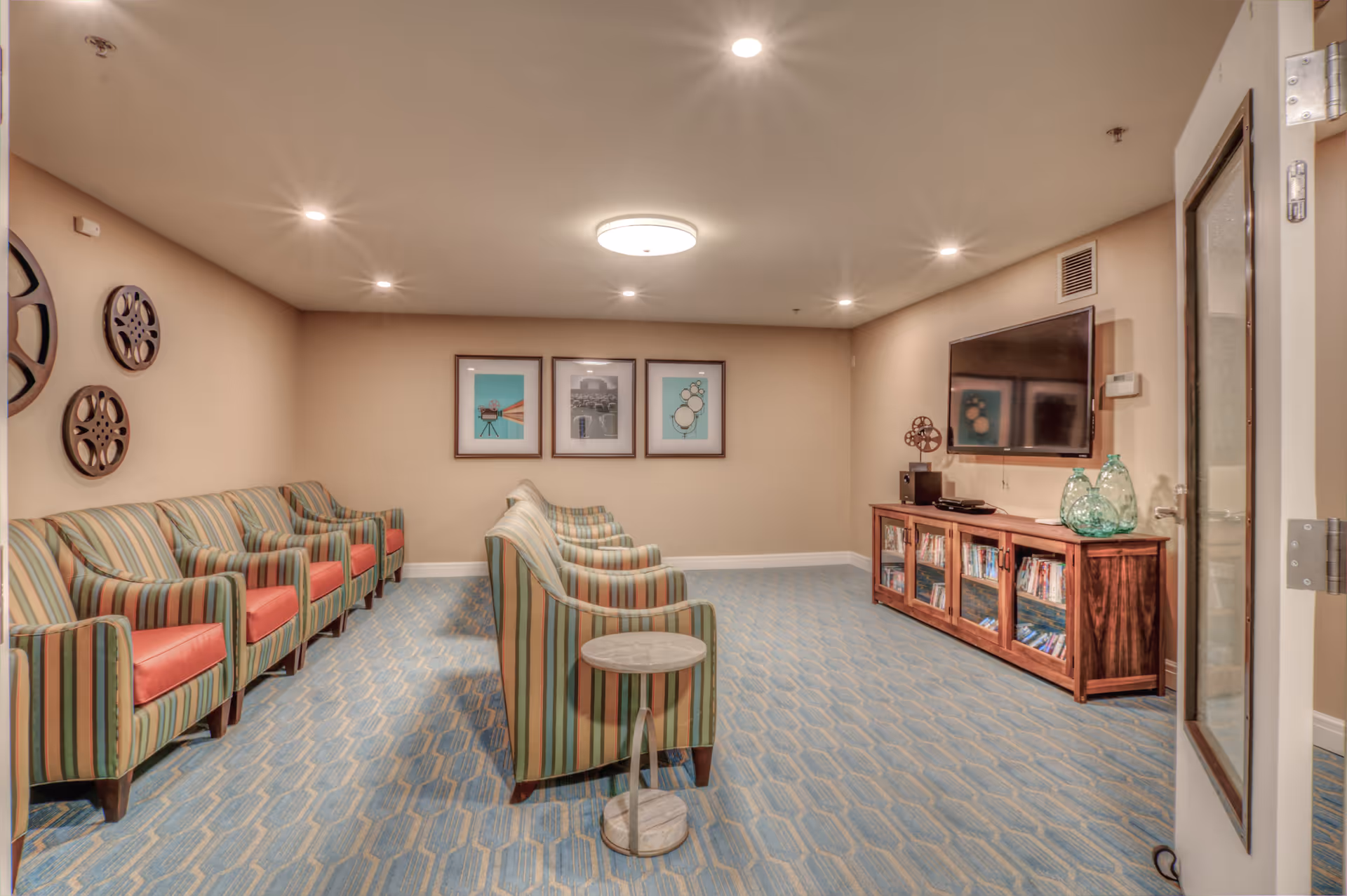 A cozy living room area in Northgate Plaza with striped armchairs arranged in two rows facing a wall-mounted flat-screen TV. The room has beige walls decorated with framed pictures and vintage film reels. A wooden cabinet below the TV holds DVDs and decorative glass vases. The floor is carpeted with a blue patterned design, and the ceiling has recessed lighting and a central light fixture.