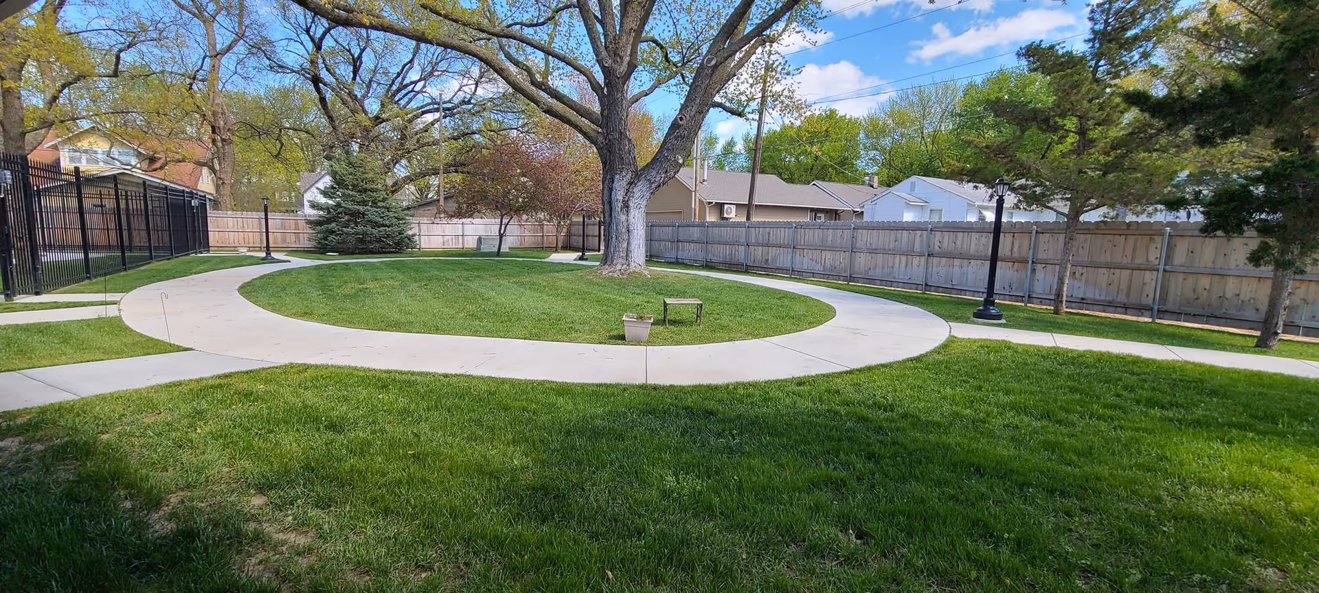 Circular concrete walkway around a grassy courtyard with a large tree, a bench, lamp posts, and a surrounding fence.