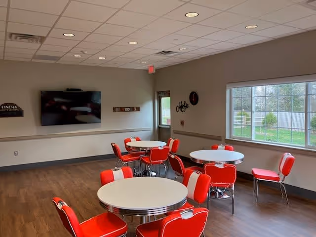 A bright common area with several round white tables surrounded by red and white retro-style chairs. The room has wooden flooring, a large window showing a fenced outdoor garden, a wall-mounted flat screen TV, and a door leading outside. The walls are light-colored with minimal decorations including a clock and a sign.
