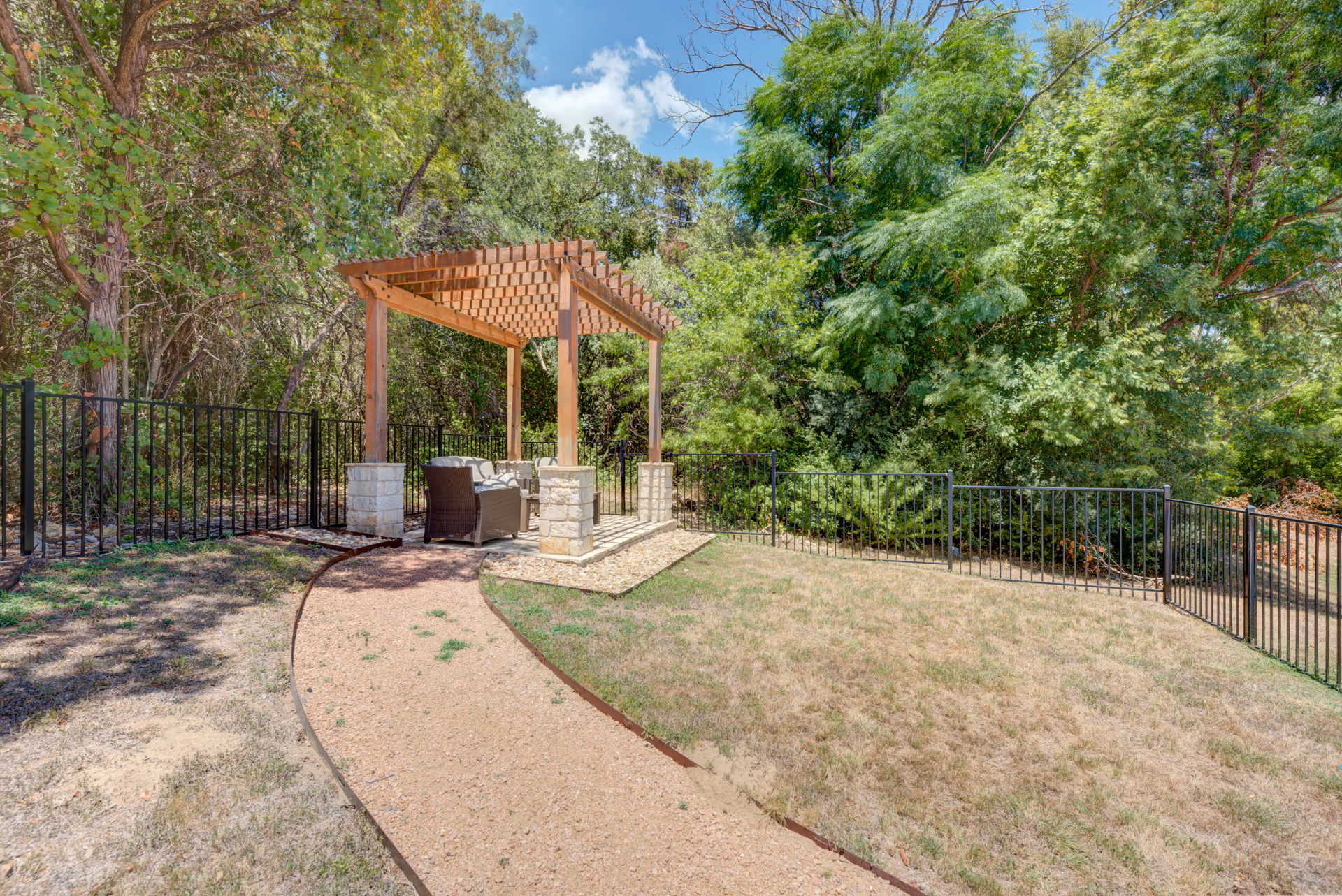 Outdoor area with a wooden pergola supported by stone pillars, containing a cushioned wicker chair and a small table. The pergola is surrounded by a black metal fence and lush green trees under a partly cloudy blue sky. A gravel pathway leads to the pergola through a grassy yard.