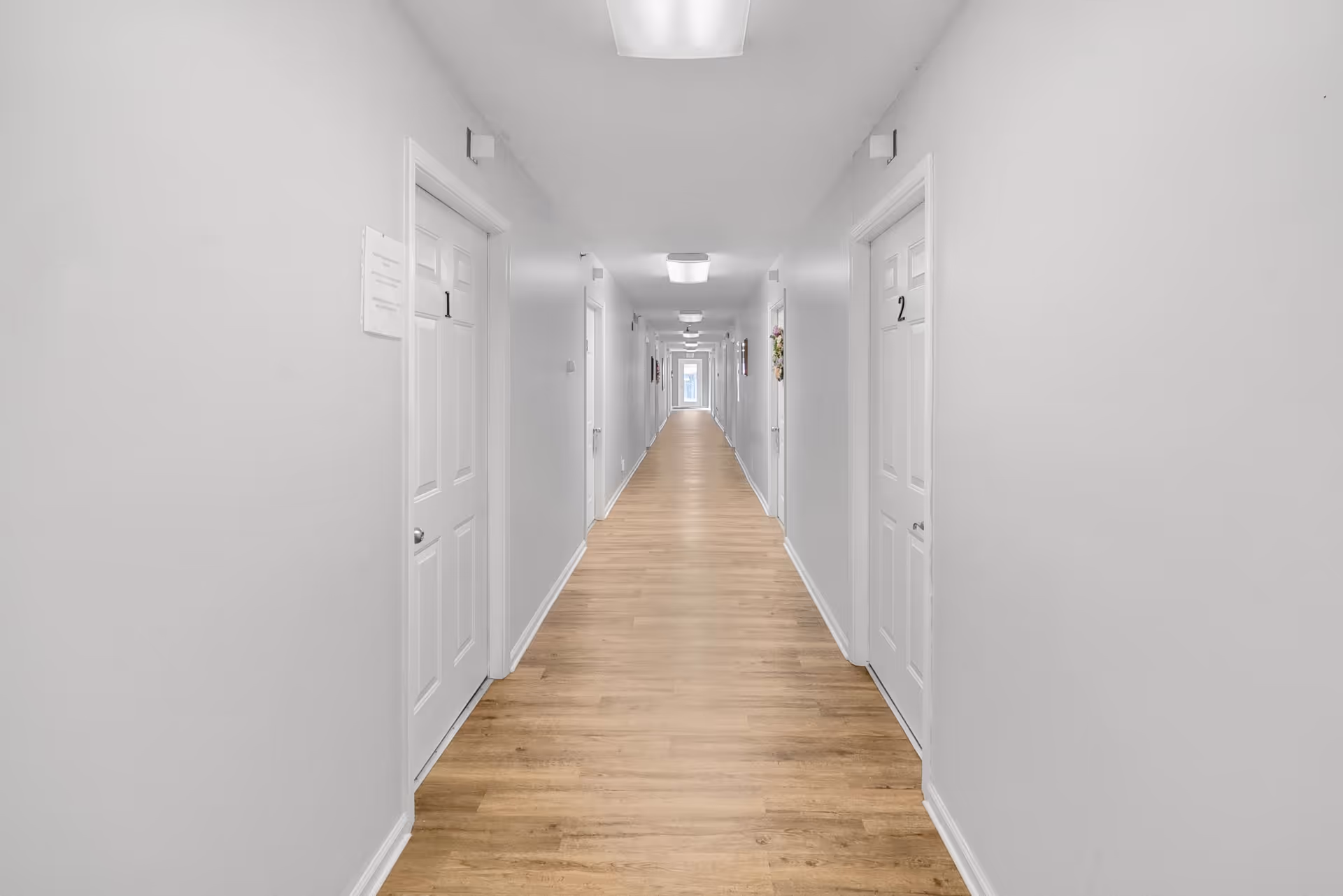 A long, narrow hallway with light wood flooring and white walls. There are multiple white doors on both sides of the hallway, each door numbered, with door 1 on the left and door 2 on the right visible in the foreground. The hallway is well-lit with ceiling lights and extends to a bright window at the far end.