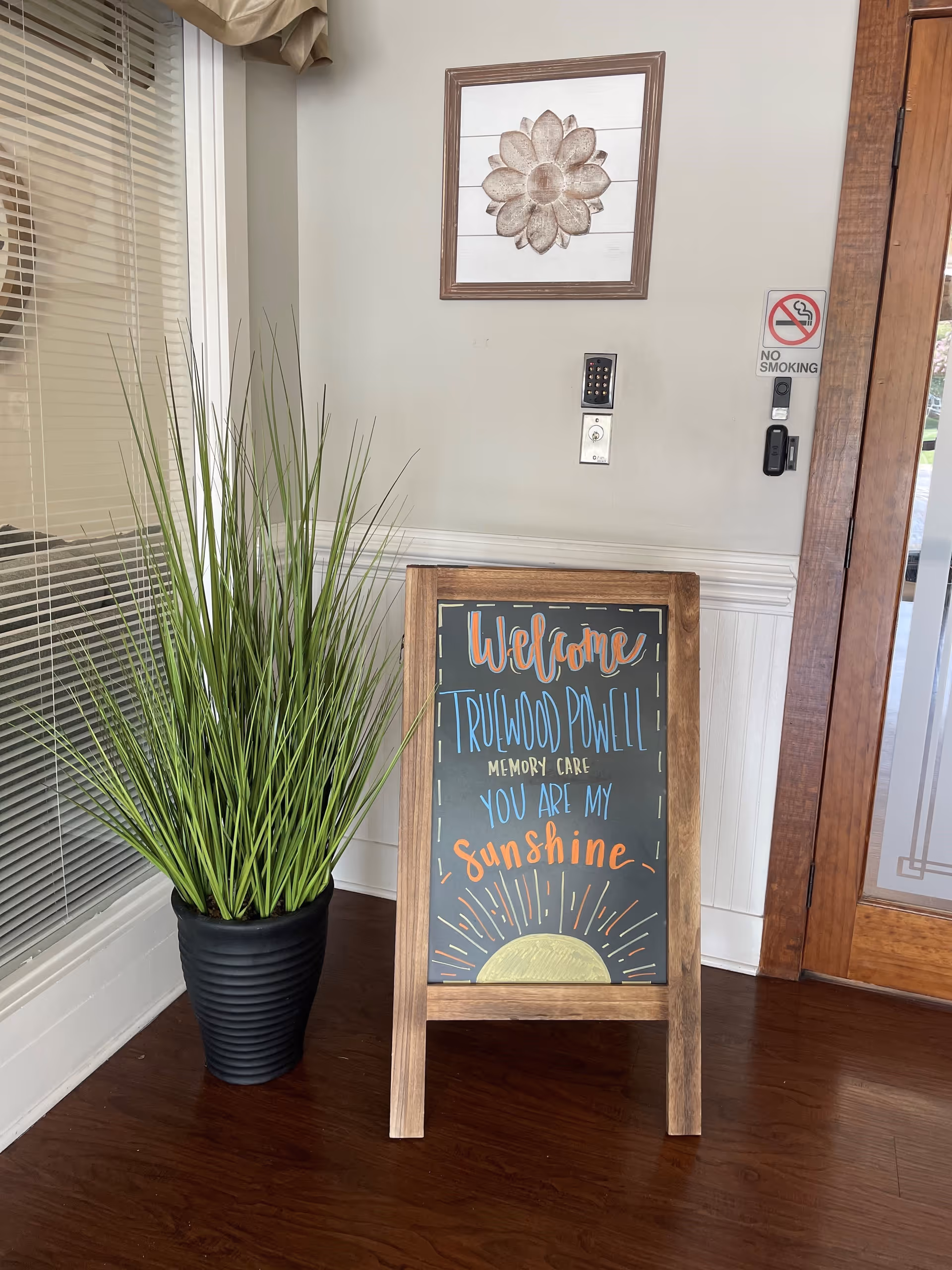 Indoor corner with a wooden floor, a tall green potted plant, and a wooden-framed chalkboard sign that reads 'Welcome Truwood Powell Memory Care You Are My Sunshine' with a drawing of a sun at the bottom. On the wall above the sign is a decorative flower artwork, a keypad, a light switch, and a no smoking sign. A wooden door with glass panels is visible on the right side.
