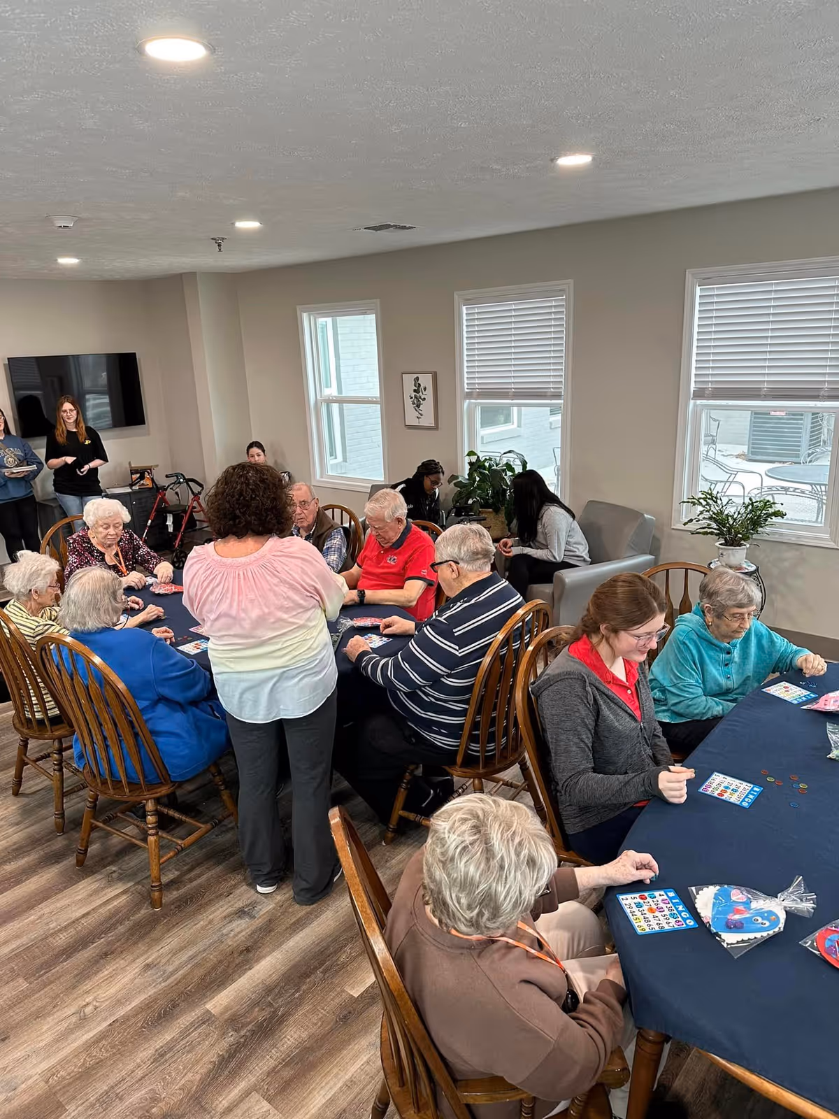 A group of elderly people and a few younger adults gathered around tables in a well-lit room playing bingo. The room has wooden floors, several windows with blinds, and some potted plants. Some people are seated while one woman stands, engaging with the group. There is a television mounted on the wall and a walker visible in the background.