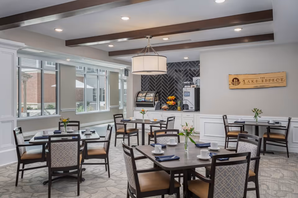 A bright and modern dining area with several tables and chairs arranged neatly. Each table has white cups, saucers, and blue napkins, along with small vases holding fresh flowers. Large windows allow natural light to fill the room. A counter at the back features a coffee machine, a display case with pastries, and a bowl of fruit. A wooden sign on the wall reads 'The Lake Effect.'