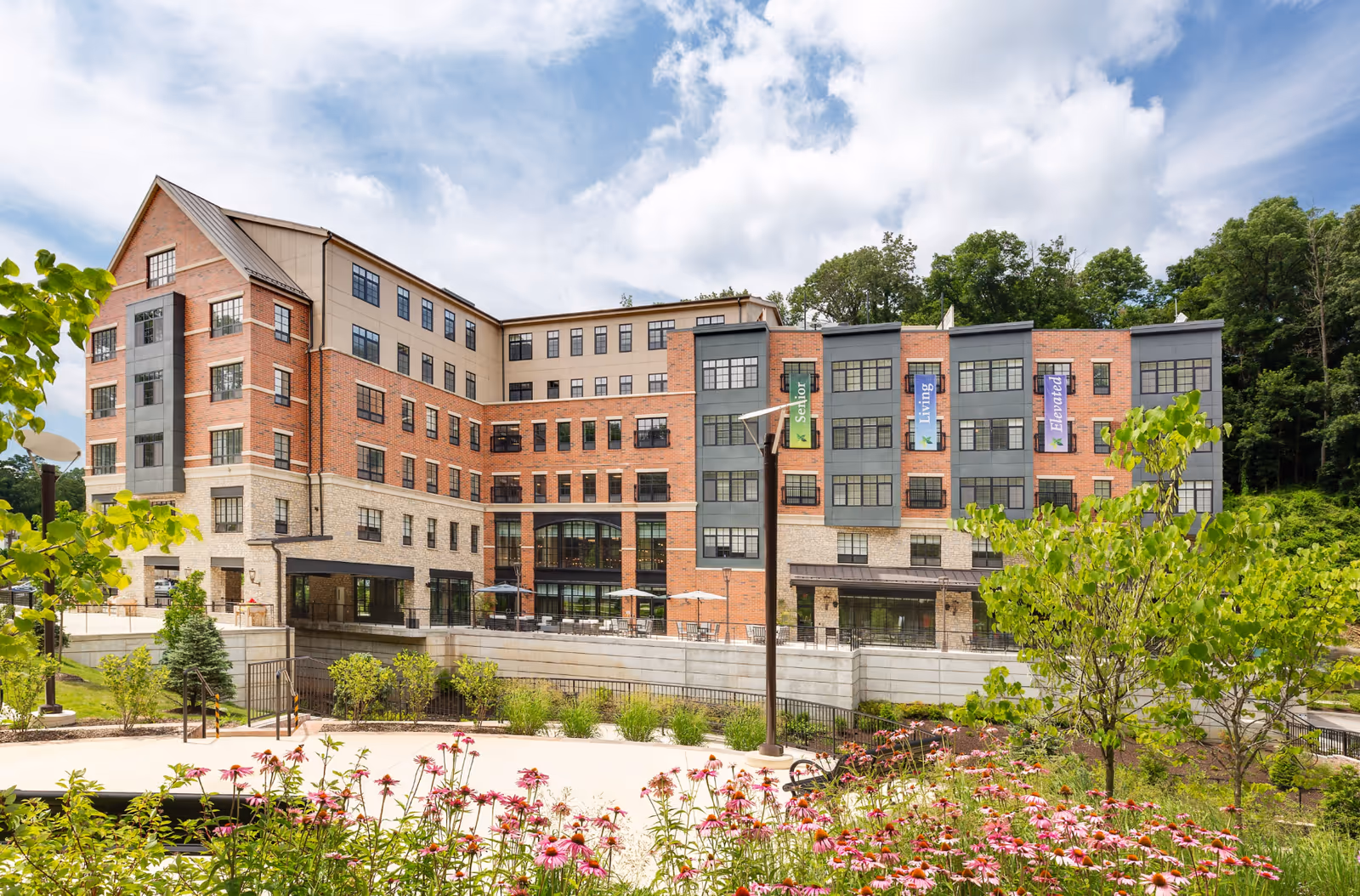 Exterior view of a multi-story senior living facility building with brick and stone facade, large windows, and banners that read 'Senior', 'Living', and 'Elevated'. The foreground features a landscaped garden with pink flowers, green shrubs, and trees under a partly cloudy sky.