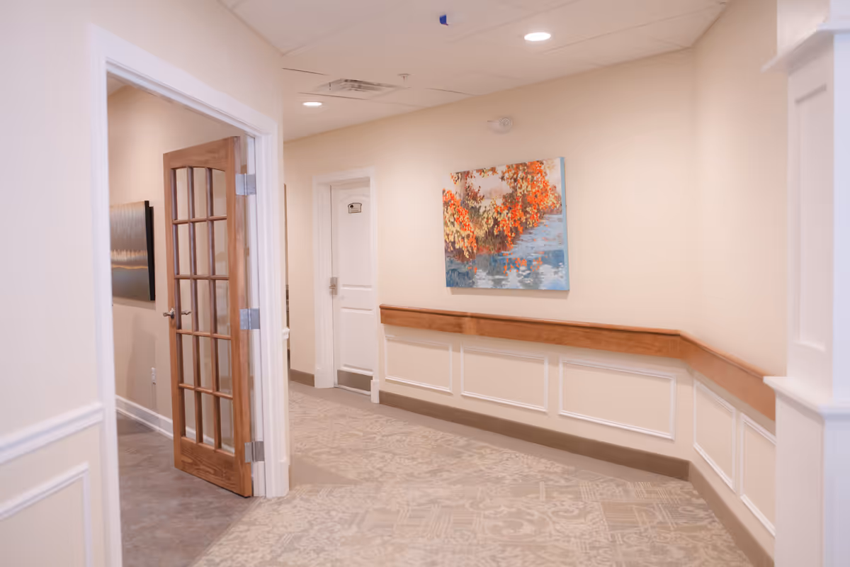 Bright assisted-living hallway with wainscoting, a wooden handrail, patterned carpet, a painting on the wall, and a glass-paneled wooden door.