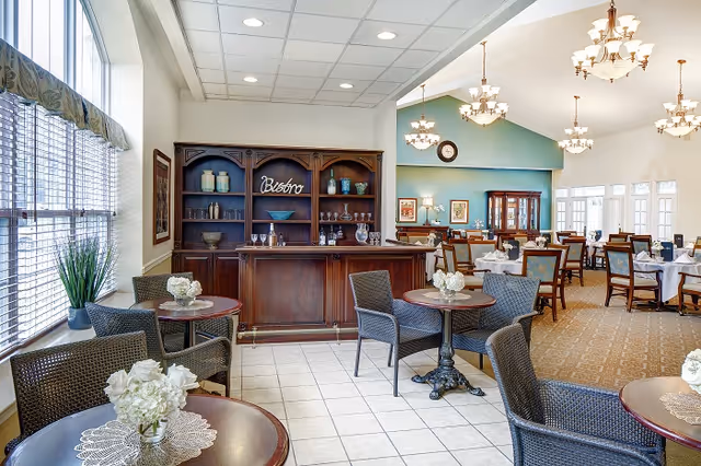 A bright dining area with multiple tables and chairs, some with white floral centerpieces. There is a wooden bistro bar with shelves holding decorative items and glassware. Large windows with blinds allow natural light to fill the room. Elegant chandeliers hang from the ceiling, and the space has a warm, inviting atmosphere.