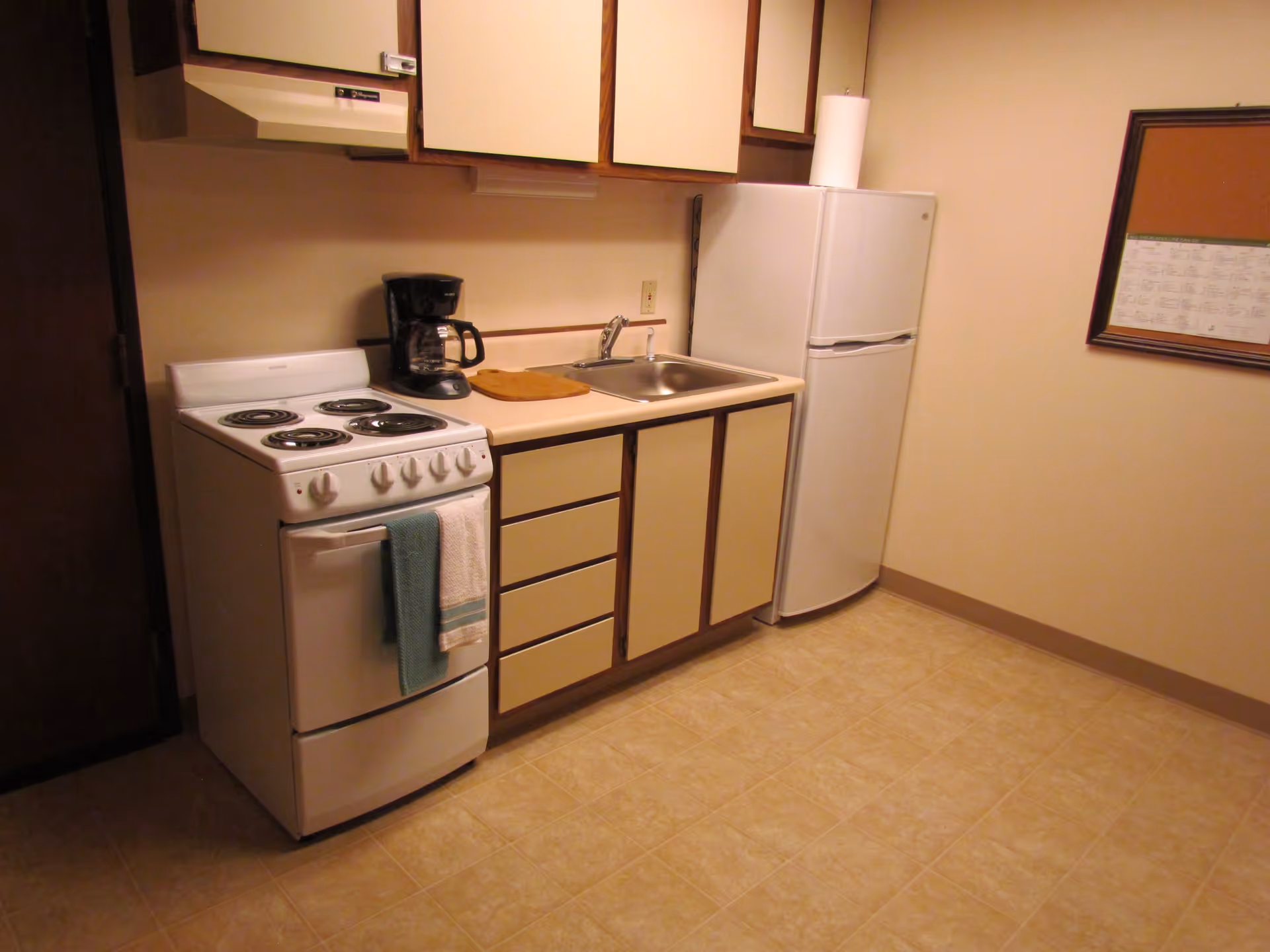 A small kitchen area with a white electric stove, a coffee maker, a sink, beige cabinets, and a white refrigerator. There is a towel hanging on the oven door and a bulletin board on the wall to the right.