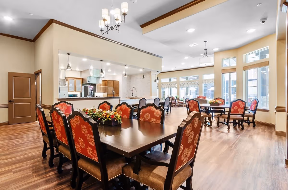 A spacious dining area with multiple dark wood tables and chairs featuring red patterned upholstery. The room has large windows allowing natural light to fill the space, and a kitchen area is visible in the background with stainless steel appliances and pendant lighting. The floor is wood, and the walls are painted in a light beige color with brown trim.