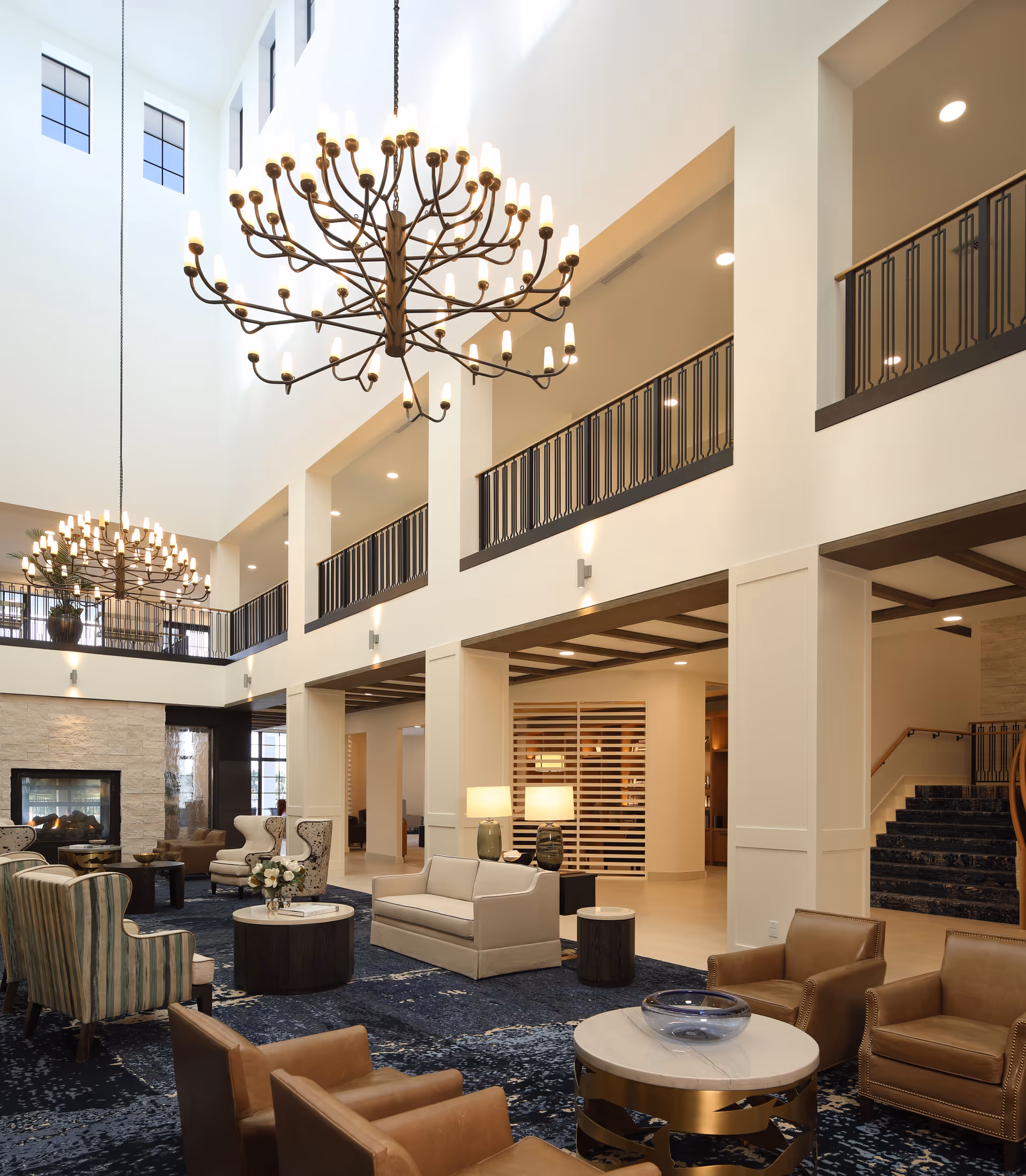 Spacious and elegant senior living facility common area with high ceilings, large chandeliers, comfortable seating including sofas and armchairs, a fireplace, and a staircase leading to an upper level with black railings. The room is well-lit with natural light from high windows and warm lamps.