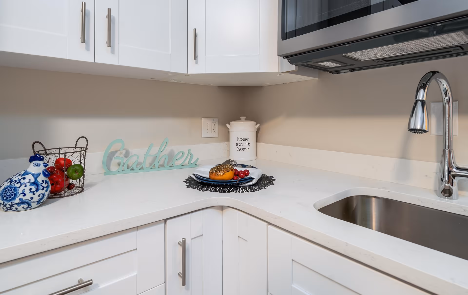 A clean and modern kitchen corner with white cabinets and a white countertop. On the counter, there is a decorative blue and white ceramic chicken, a wire basket with red and green apples, a light blue decorative sign that says 'Gather', a white container labeled 'home sweet home', and a plate with a small pumpkin and cherries on a black doily. A stainless steel sink and a chrome faucet are visible on the right side.