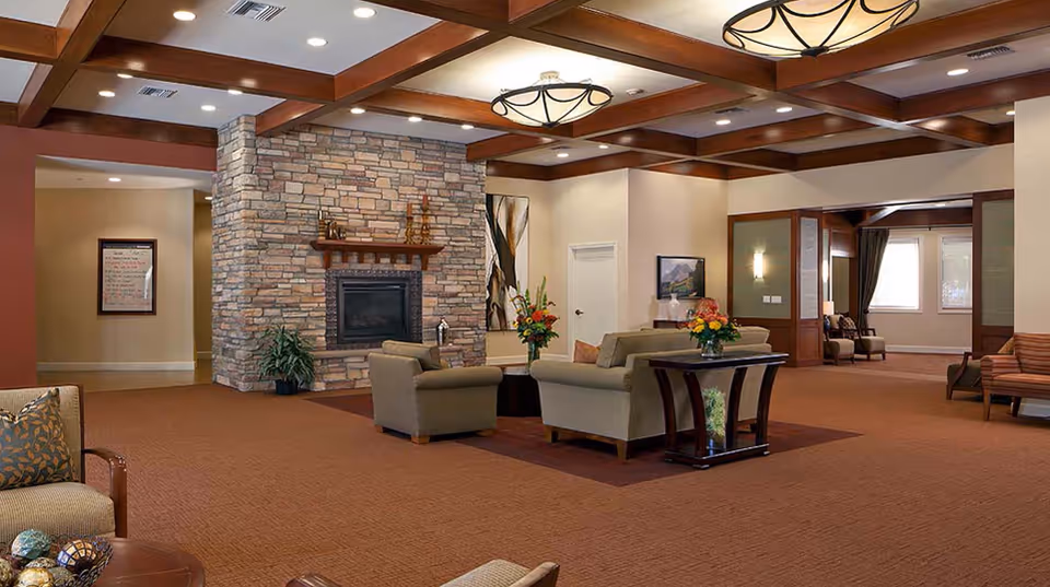 Spacious senior living lounge with grouped sofas and chairs arranged around a stone fireplace under coffered ceilings.