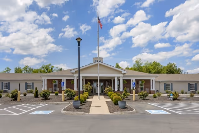 Front entrance of a single-story senior living facility with a flagpole, walkway, landscaping, and accessible parking.
