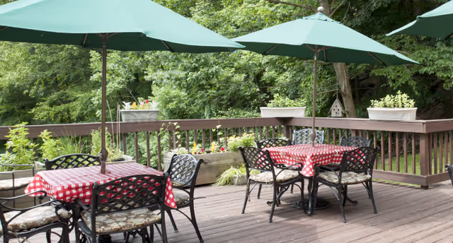 Outdoor wooden deck with two tables covered in red and white checkered tablecloths, each table shaded by a large green umbrella. Black metal chairs with patterned cushions surround the tables. The deck is bordered by a wooden railing with planter boxes containing flowers and greenery, and lush green trees are visible in the background.