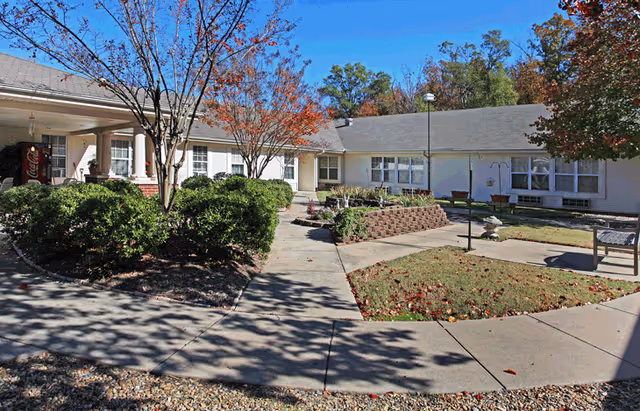 Outdoor courtyard area at Willow Grove of Maumelle featuring a paved walkway, landscaped bushes, trees with autumn leaves, benches, and a raised garden bed in front of a single-story building with multiple windows under a clear blue sky.