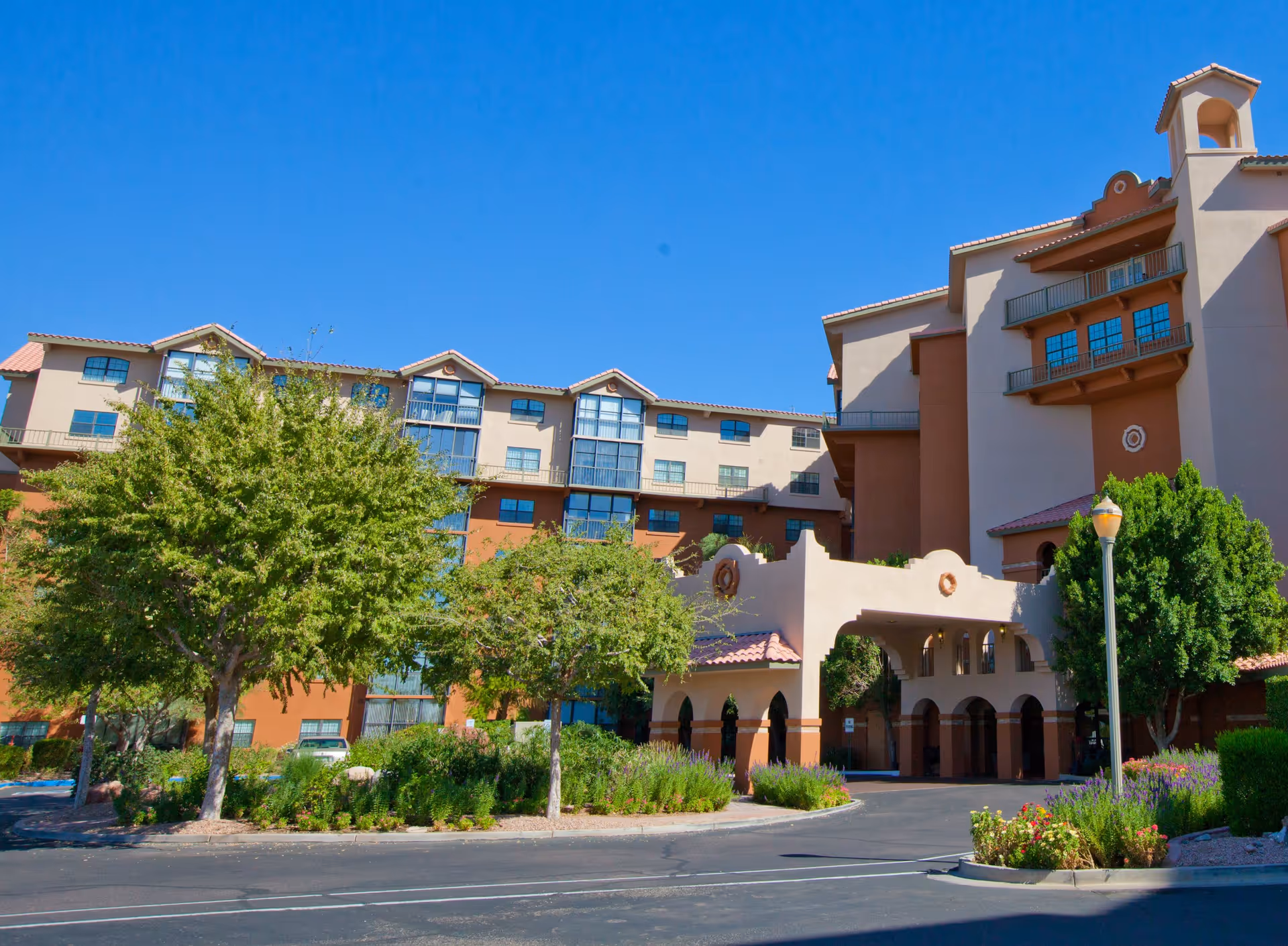 Exterior view of a large multi-story building with a southwestern architectural style, featuring beige and terracotta colors, arched entryways, balconies, and surrounded by trees and landscaped greenery under a clear blue sky.