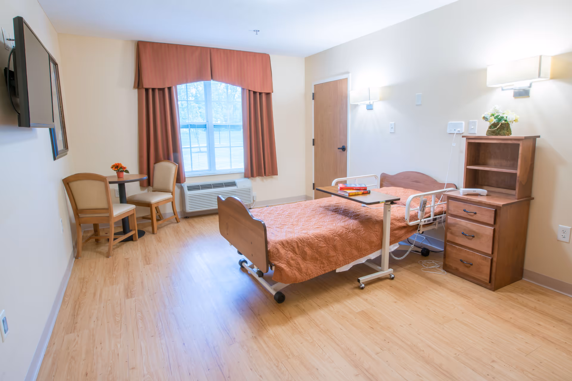 A senior living facility bedroom with a single hospital-style bed covered with a brown quilt, a wooden bedside cabinet with a small flower arrangement, a small table with two chairs near a window with brown curtains, and a wall-mounted TV.