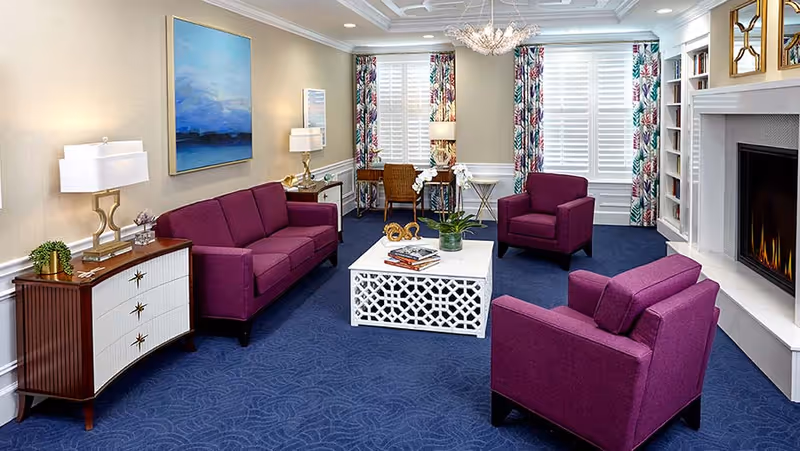 A cozy living room with purple upholstered sofa and armchairs arranged around a white decorative coffee table. The room features a blue carpet, a fireplace on the right, two windows with colorful patterned curtains, a wooden sideboard with two lamps, and a desk with a chair near the windows.