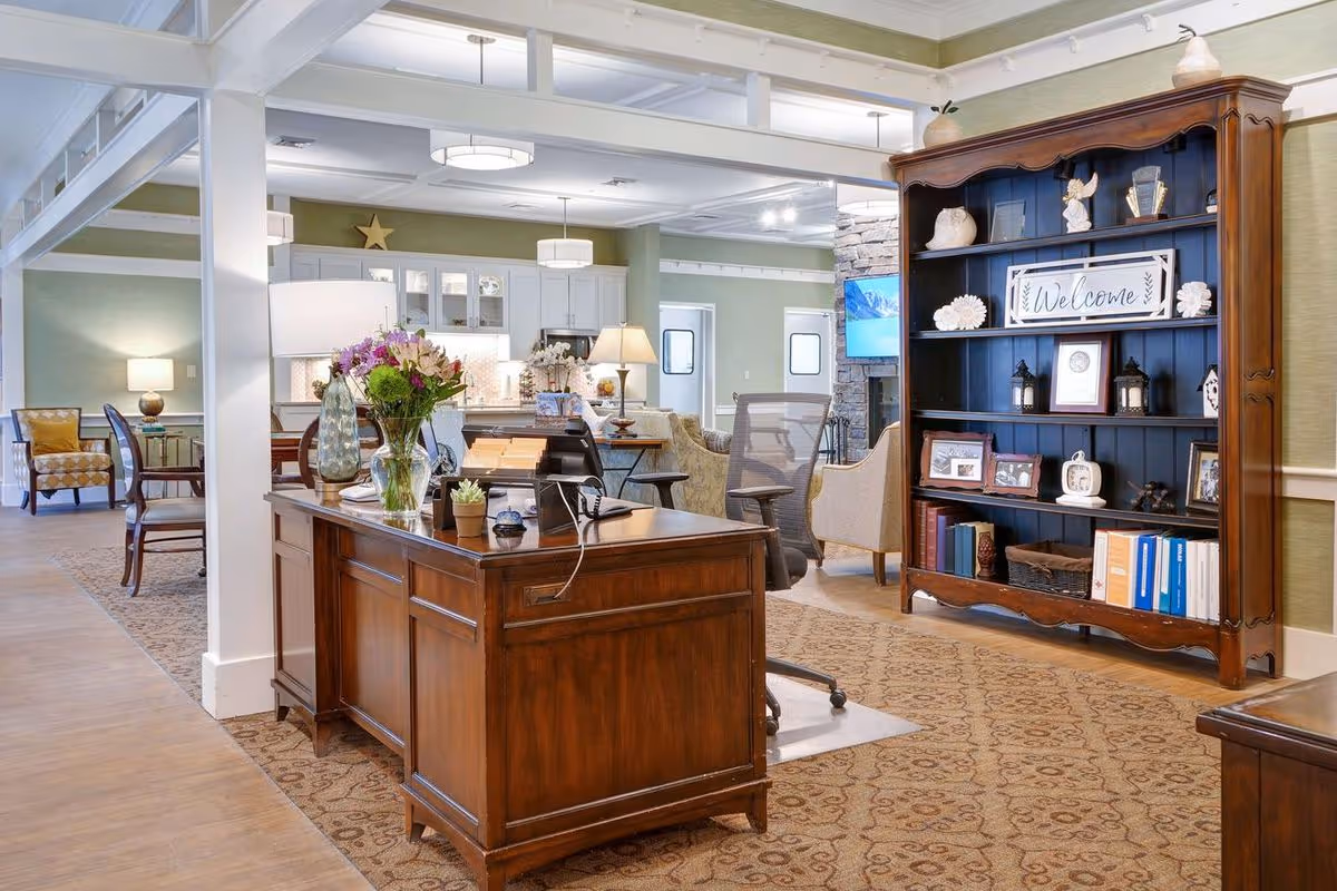A bright and welcoming interior space of a senior living facility featuring a wooden reception desk with flowers and office supplies, a large wooden bookshelf with decorative items and a 'Welcome' sign, comfortable seating areas with chairs and lamps, and a kitchen area in the background.
