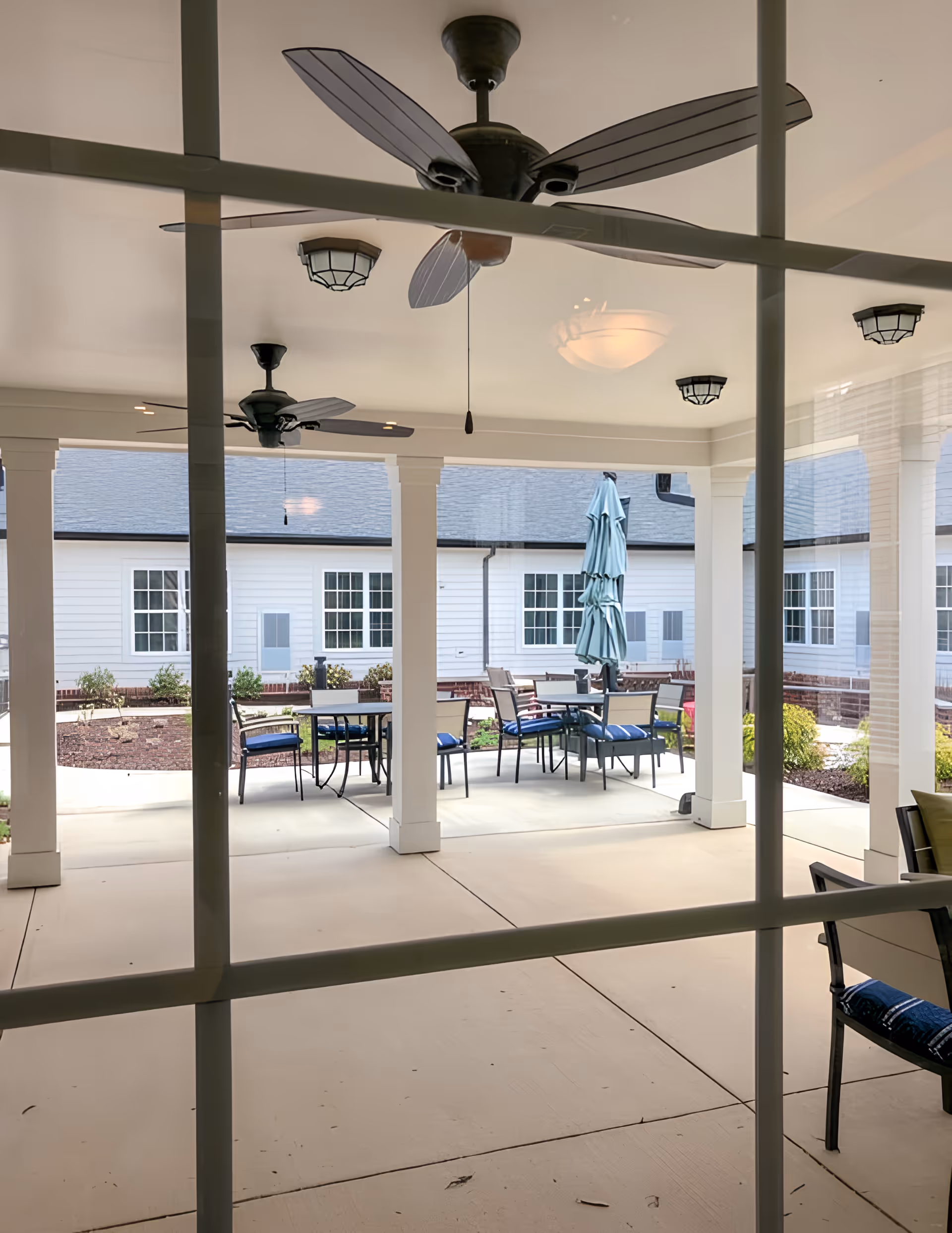 View through window panes of a covered outdoor patio with ceiling fans, tables, chairs, and a closed umbrella in a courtyard.