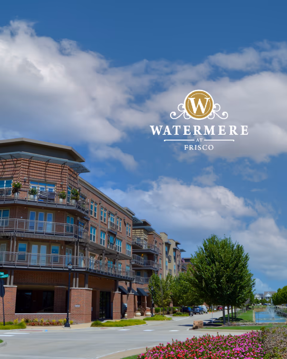 Exterior view of a multi-story brick and beige building with balconies, surrounded by trees, a flower bed, and a water fountain under a partly cloudy blue sky. The logo 'Watermere at Frisco' is overlaid in the sky area.