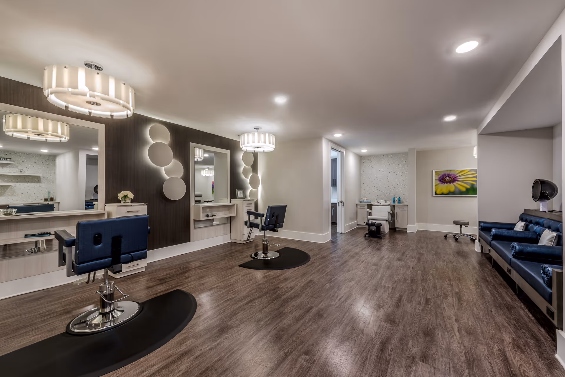 Interior view of a modern salon area in a senior living facility with two styling chairs in front of large mirrors on the left, a salon chair and counter with hair products in the back, and a blue cushioned seating area with a hair dryer on the right. The room has wood flooring, recessed lighting, and decorative wall elements.