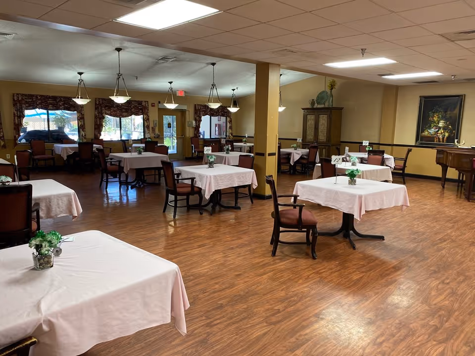 A spacious dining room with multiple square tables covered with white tablecloths, each table having a small flower arrangement and a table number. The room has wooden flooring, beige walls, hanging ceiling lights, and windows with patterned curtains. There is a wooden cabinet and a framed painting on the far wall.