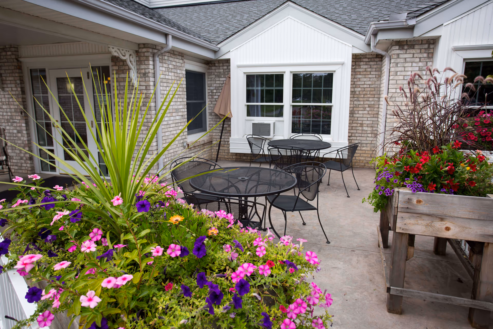 Outdoor patio area at Ecumen Centennial House featuring black metal tables and chairs, surrounded by vibrant flower planters with pink, purple, and red flowers. The building exterior is made of light-colored brick with white trim and windows, one of which has an air conditioning unit.