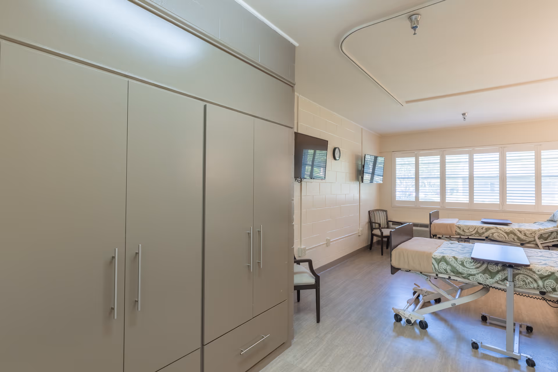 A bright shared resident room with two adjustable beds, wall-mounted TVs, large storage cabinets and chairs by a row of windows.