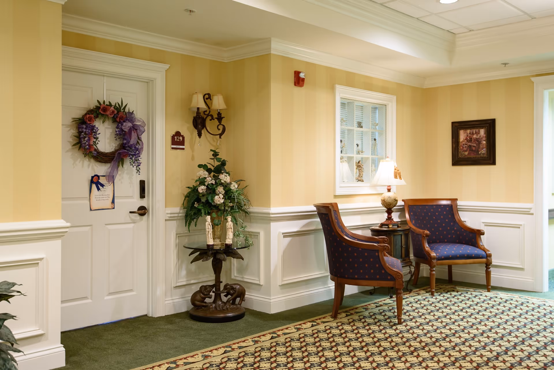 Hallway seating area with two upholstered chairs, a side table and lamp, decorative plants and a wreath on a door.