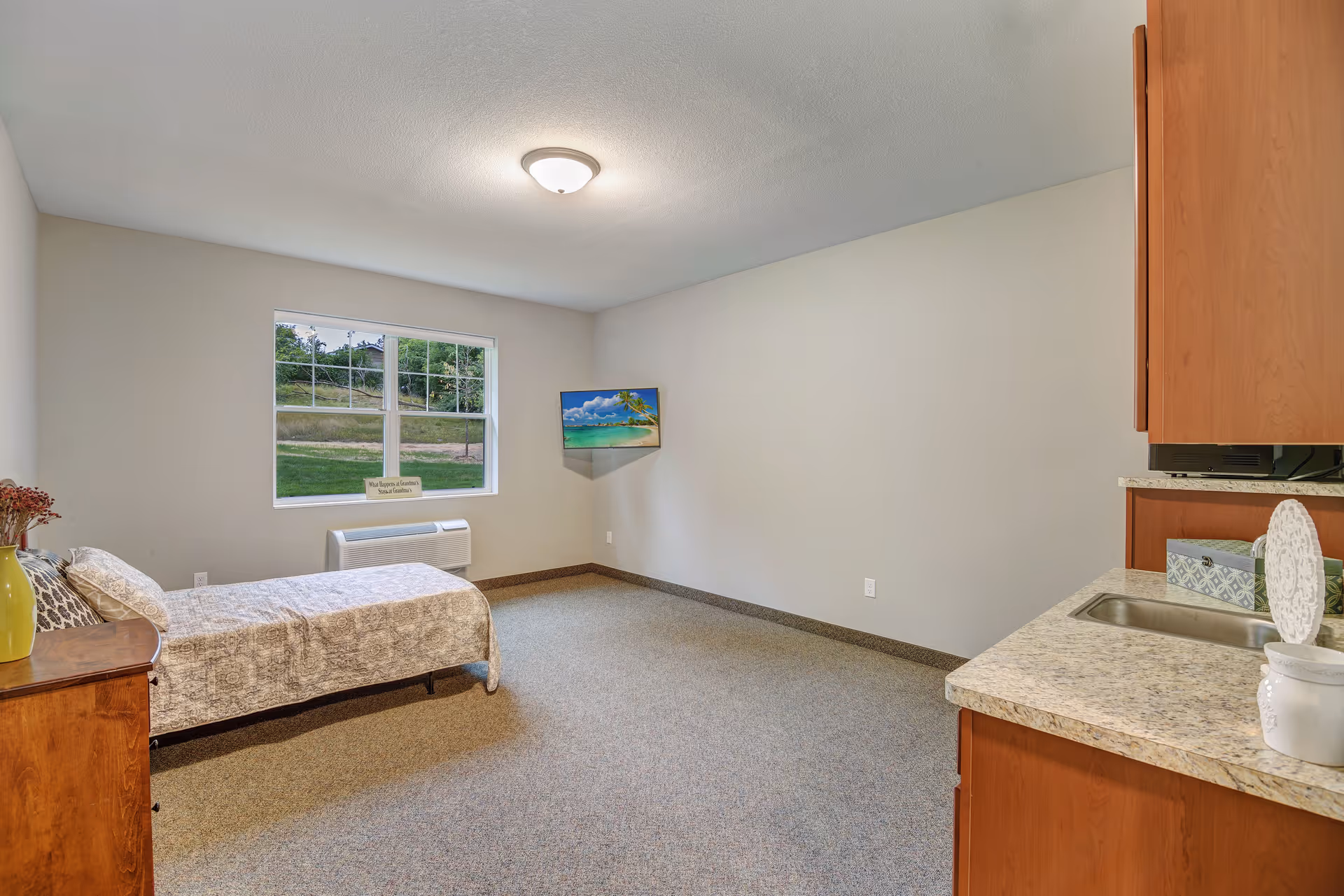A senior living facility bedroom with a single bed covered in a patterned bedspread, a wooden nightstand with a yellow vase and dried flowers, a window showing greenery outside, a wall-mounted TV displaying a beach scene, and a kitchenette area with a sink and cabinets.