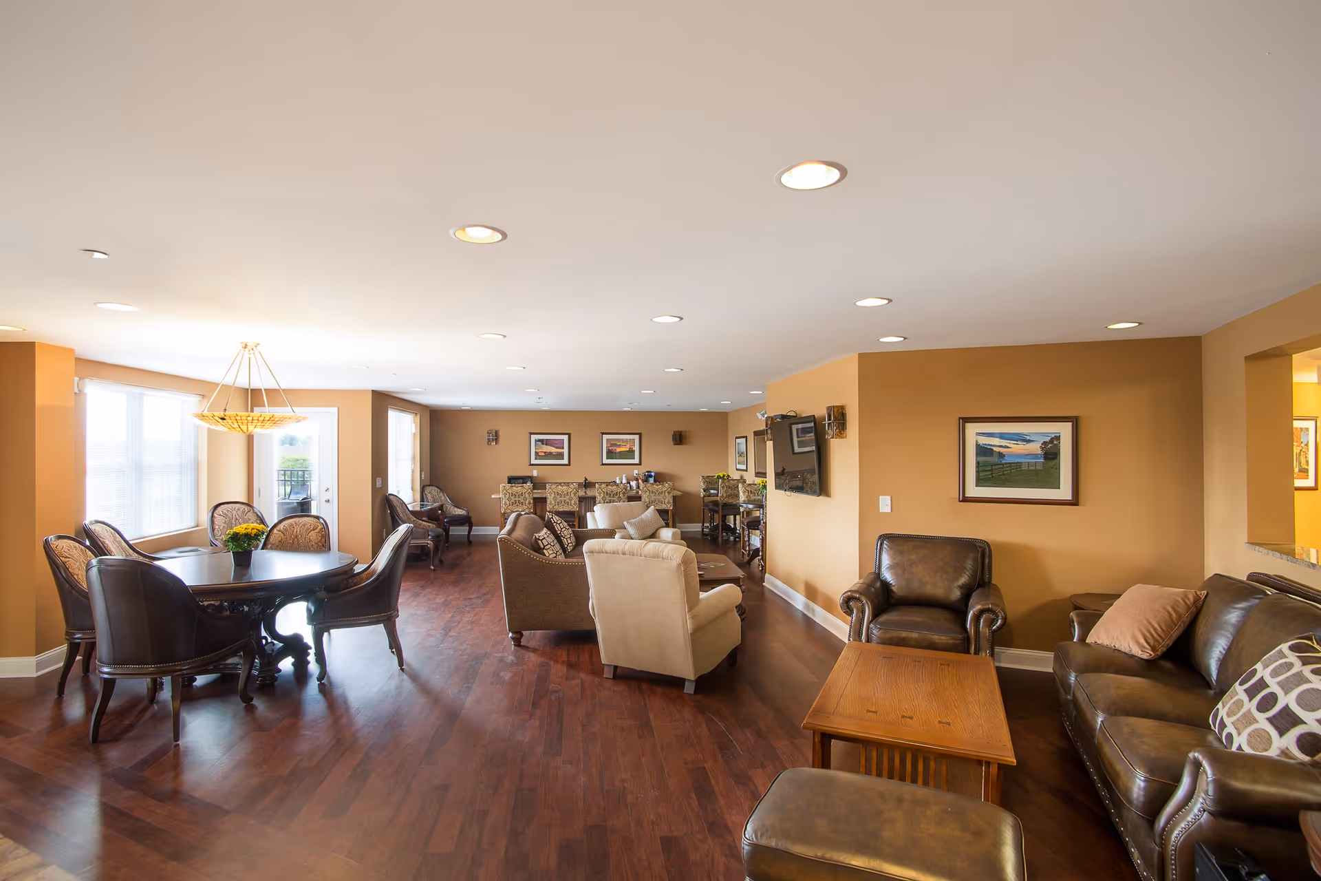 A spacious and well-lit common living area with wooden flooring, featuring a round dining table with six chairs on the left, multiple armchairs and sofas arranged around a wooden coffee table on the right, and a wall-mounted TV. The walls are painted a warm beige color and decorated with framed pictures. Large windows and a glass door allow natural light to fill the room.