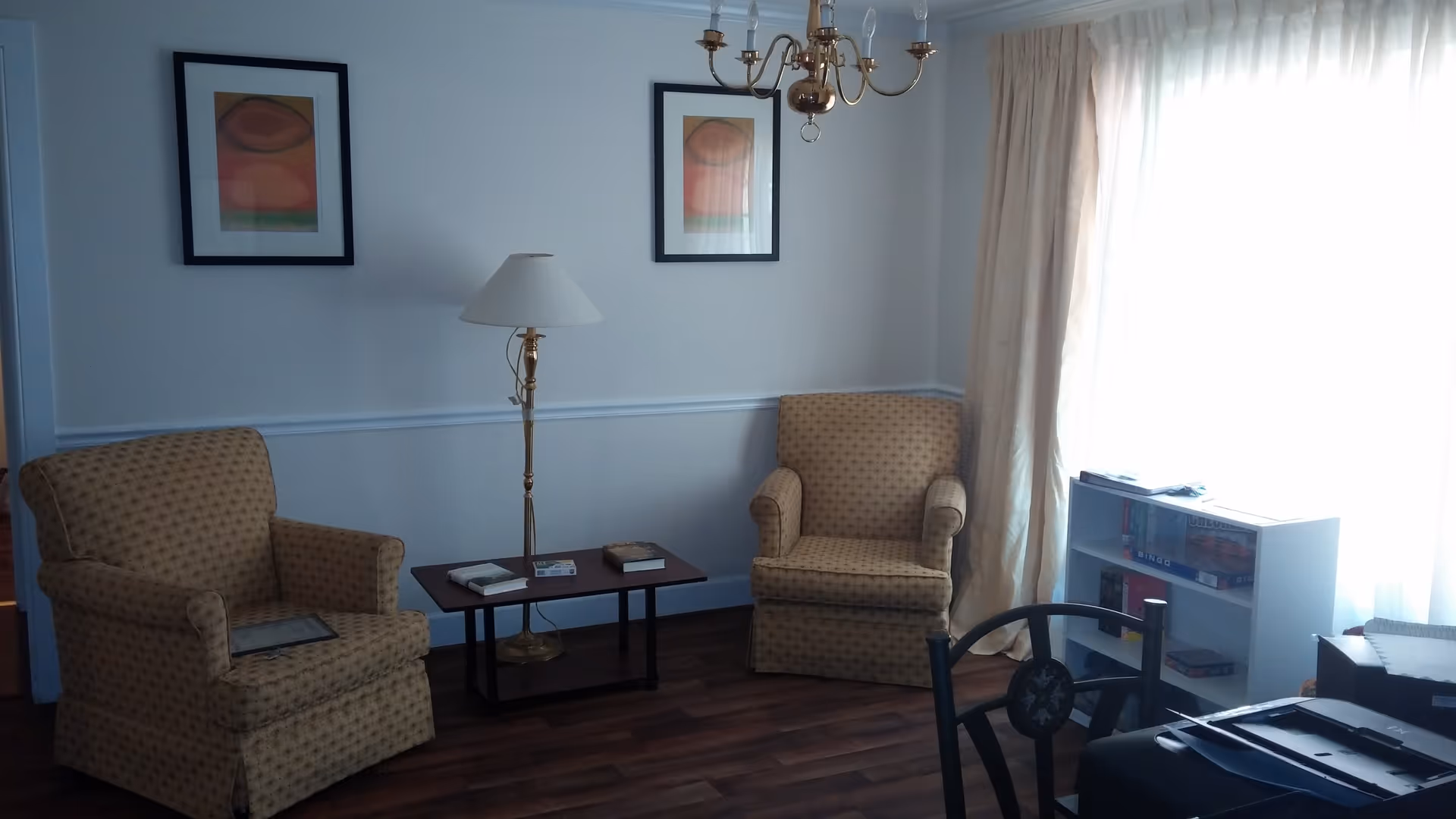 A cozy sitting area with two patterned armchairs facing each other, a small wooden table with books and a lamp between them, two framed abstract paintings on the wall, a chandelier hanging from the ceiling, and a window with sheer curtains letting in bright light. There is also a white bookshelf with board games and books near the window.