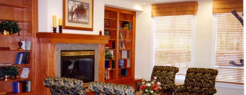 Cozy living room area with floral upholstered chairs arranged around a coffee table with a flower arrangement. The room features a wooden fireplace with a framed picture above it, built-in wooden bookshelves filled with books and decorative items, and windows with wooden blinds letting in natural light.