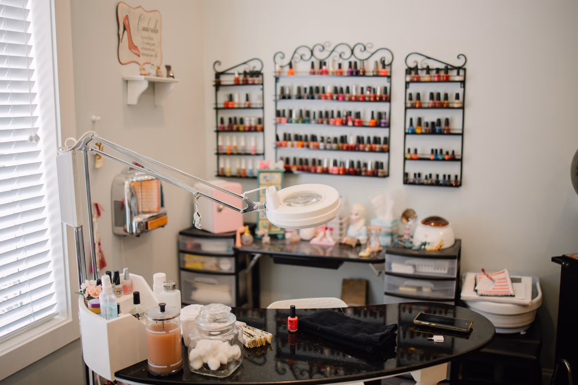 A manicure station in a room with a black countertop table holding nail care items including cotton balls, nail polish remover, and a red nail polish bottle. Behind the table, there are three wall-mounted racks filled with various colors of nail polish bottles. The room has a window with white blinds on the left side and a small shelf with decorative items on the wall.