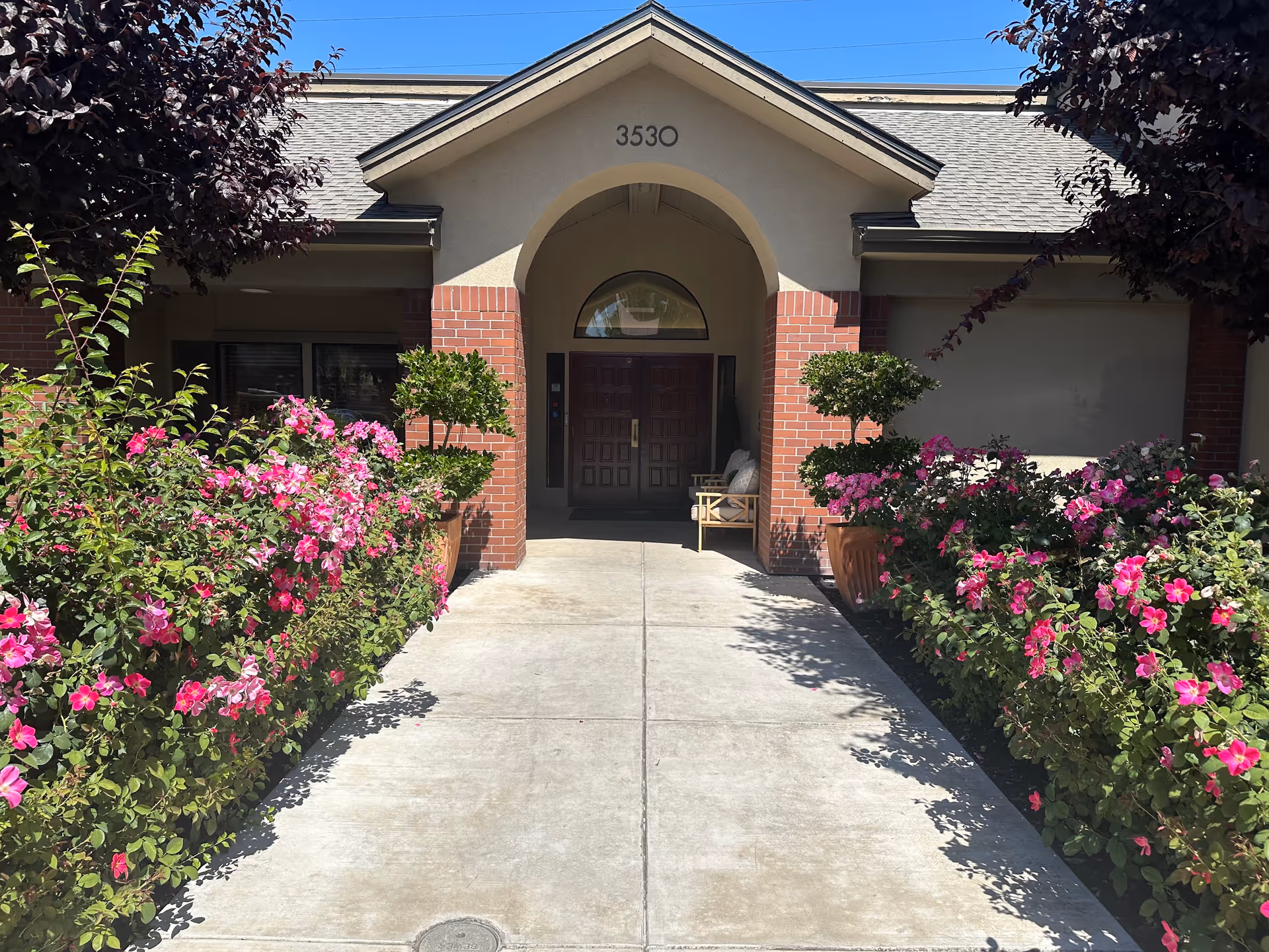 Front entrance of a building with the address number 3530 above an arched doorway. The entrance is flanked by brick columns and potted plants with trimmed greenery. There are vibrant pink flowers lining both sides of the concrete walkway leading to the door. The sky is clear and blue.