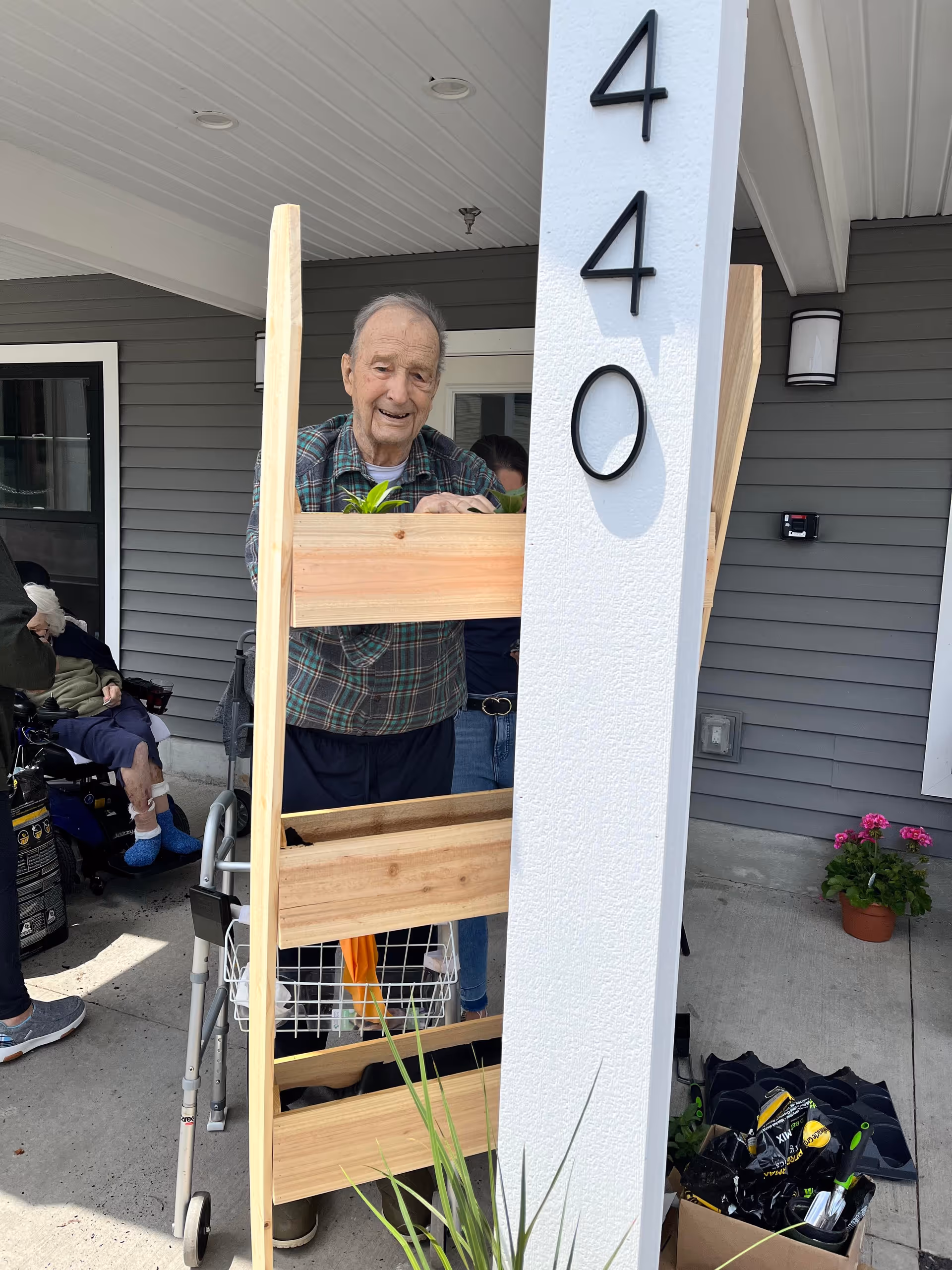 An elderly man standing outside a building with the number 440 on a white pillar. He is smiling and holding a wooden planter box. In the background, another elderly person is seated in a wheelchair, and there are some plants and gardening supplies nearby.