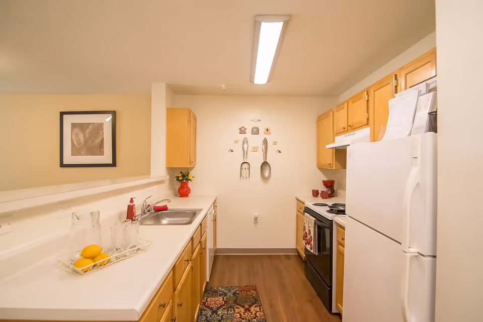 A compact kitchen with wooden cabinets, white countertops, and a white refrigerator. The kitchen features a stainless steel sink, an electric stove with a range hood, and decorative wall art of a fork and spoon. There is a tray with lemons and glasses on the counter, a red vase with flowers, and a patterned rug on the wooden floor.