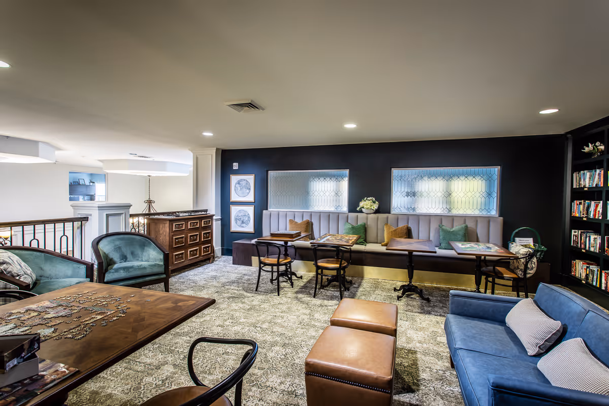 A cozy common area with a mix of seating options including a blue sofa with pillows, two brown ottomans, green armchairs, and a long cushioned bench along a dark wall. Several small tables are placed in front of the bench, some with puzzles on them. The room has a carpeted floor, bookshelves filled with books, and decorative framed artwork on the walls.