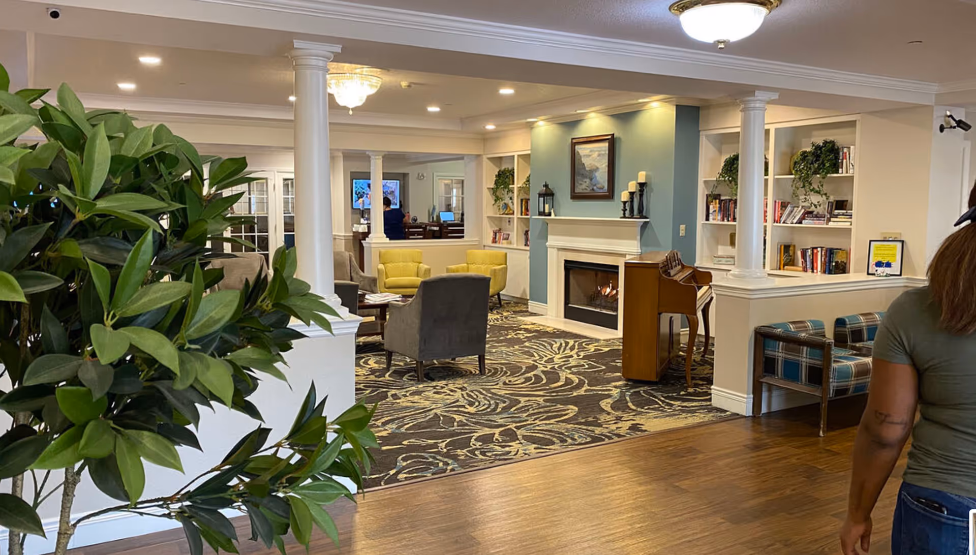 Interior view of a senior living facility lounge area with a patterned carpet, several chairs including yellow and gray armchairs, a fireplace with candles on the mantel, built-in bookshelves with books and plants, a piano, and a large green plant in the foreground. A person is partially visible walking on the right side of the image.