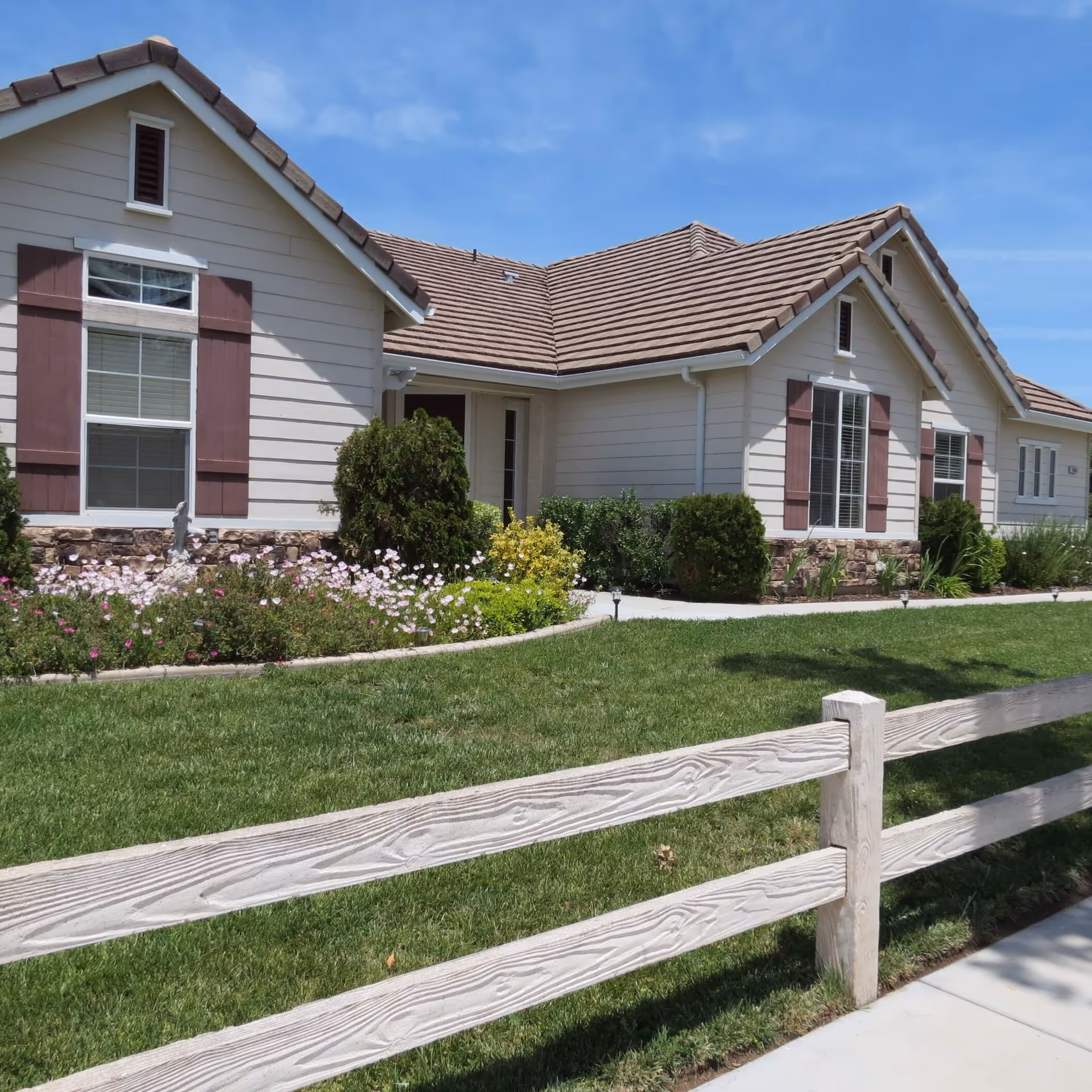 Single-story light-colored building with brown shutters, landscaped flower beds, a green lawn and a low wooden fence under a blue sky.