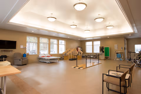 A spacious rehabilitation room with large windows letting in natural light. The room contains various physical therapy equipment including parallel bars, a therapy bed with pillows, exercise machines, chairs, and a small set of stairs with handrails. The ceiling has recessed lighting fixtures and the walls are painted in a light neutral color.