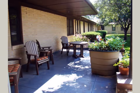 Outdoor patio area with several wooden chairs with cushions and a small table, adjacent to a brick building. Large potted plants and greenery are visible along the patio, with trees and grass in the background under a sunny sky.